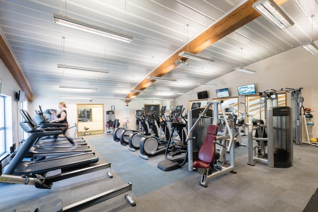 Training machines in a gym. A member can be seen moving on a treadmill in the background